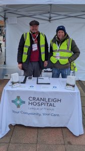 Two people standing behind a table draped in a white tablecloth that says Cranleigh Hospital
