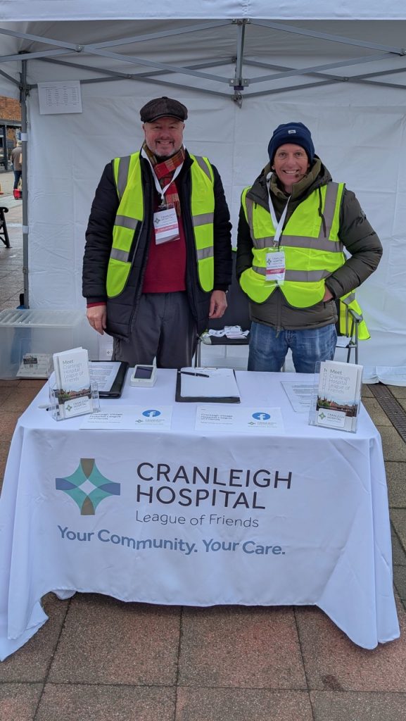 Two people standing behind a table draped in a white tablecloth that says Cranleigh Hospital