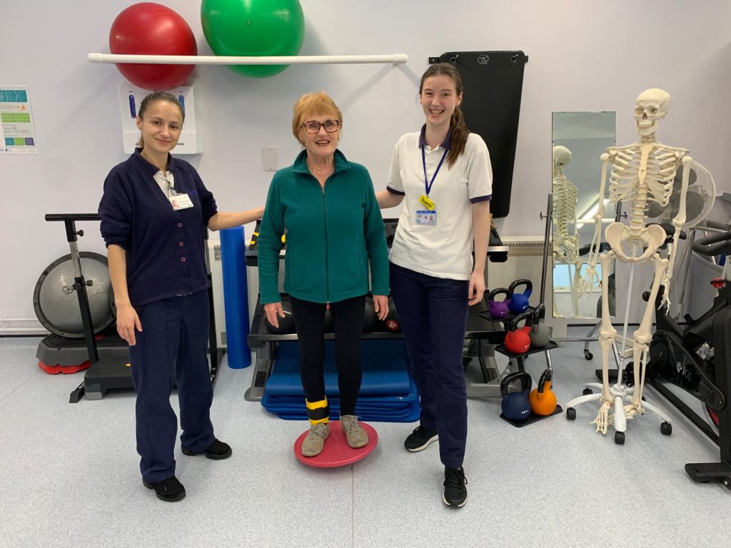 Lady balancing on a wobble board. Physiotherapists on each side help to support her.
