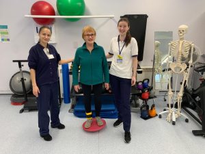 Lady balancing on a wobble board. Physiotherapists on each side help to support her.
