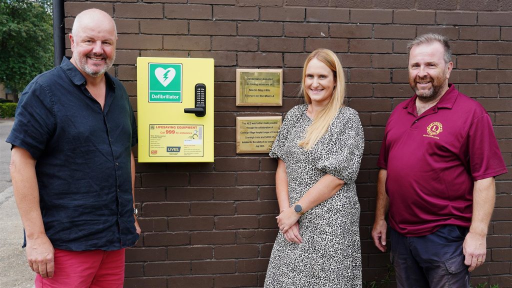 Three people standing next to a wall mounted defibrillator with two brass plaques to the right.