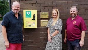 Three people standing next to a wall mounted defibrillator with two brass plaques to the right.